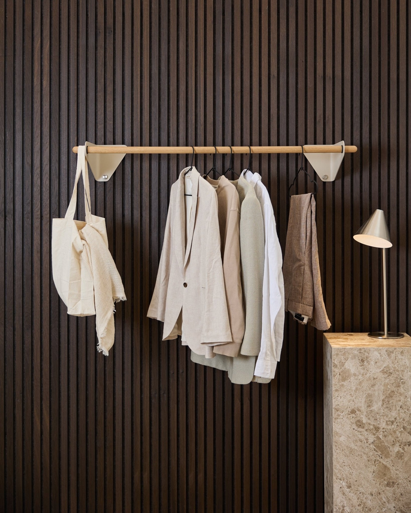 Een beige WoodUpp Linn kapstok (115,9 cm) draagt jassen, een wit overhemd en een crèmekleurige tote bag, voor een donkerhouten Akupanel Akoestisch paneel en een marmeren lamp.